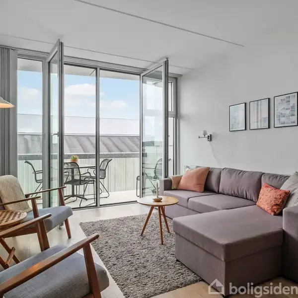 En stue med en grå hjørnesofa og puder, står foran gulv-til-loft-vinduer, der åbner ud til en altan med bord og stole. På væggen hænger tre billeder.