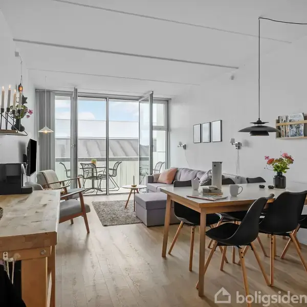Stue med spisebord, stole og sofa, i lys og moderne indretning. Store vinduer leder ud til en balkon med udsigt. Møbleret med lamper og dekorationer på hylder.