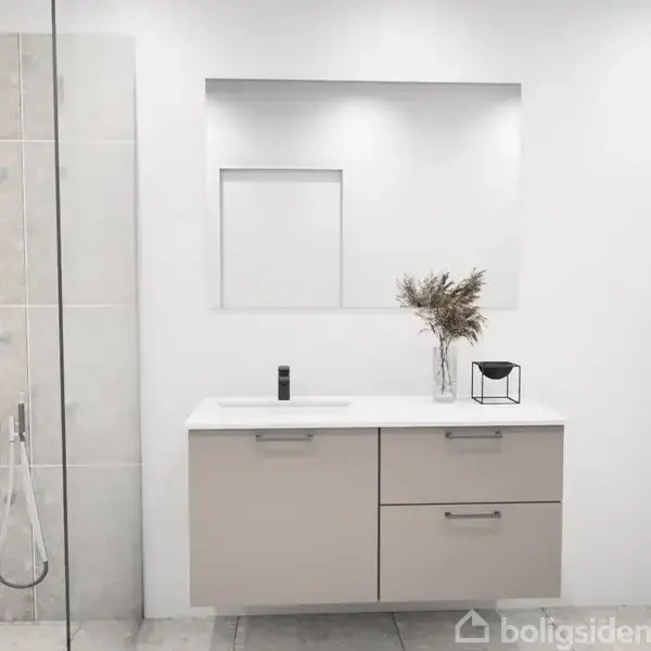 Badeværelse med en hvid vask på et beige skab. Et spejl hænger over vasken. Brusekabine til venstre. En vase med tørrede planter står på skabet.