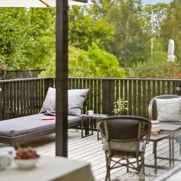 En træterrasse med en liggestol og et bord under en parasol; omgivet af grønne træer og buske. Der står også en stol og en plante ved siden af bordet.