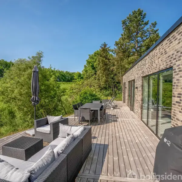 Terrasse med havemøbler, herunder sofaer og spisebord, står på en stor trædæk. Bygningen med murstensfacade er på højre side, omgivet af grønne træer og buske under en klar blå himmel.