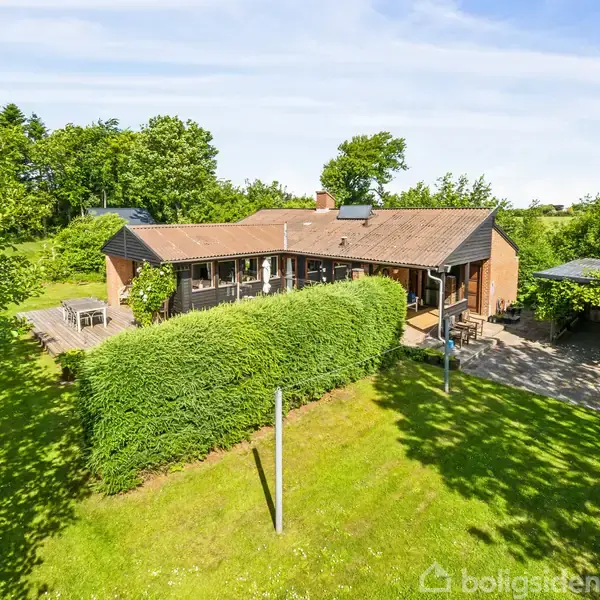Et rødt murstenshus ligger omgivet af en frodig have med græsplæne og buske. Huset har store vinduer og en terrasse. Træer omkranser ejendommen under en klar himmel.