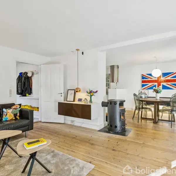 En moderne stue med sort sofa indeholder et rundt sofabord og brændeovn. I baggrunden ses et spisebord med stole og et britisk flag på væggen. Vægge prydes af kunstværker.