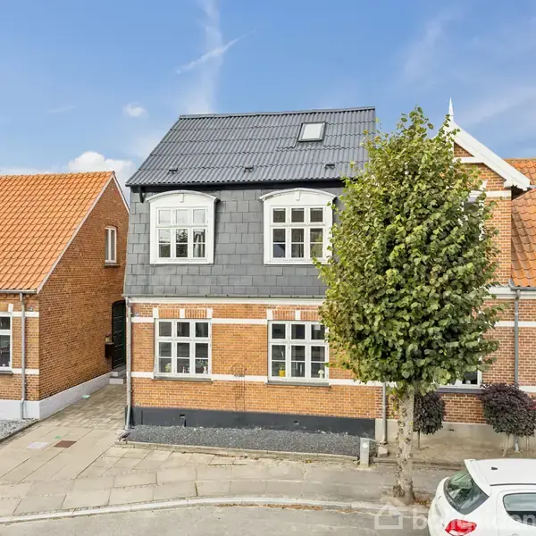Hus med røde mursten og gråt tag står på en rolig vej. Nabohusene har røde tage. En hvid bil holder parkeret ved fortovet. Der står et træ foran huset.