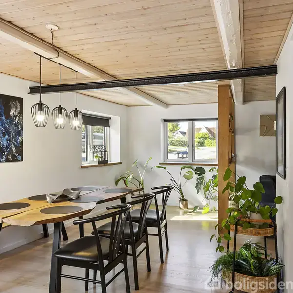 Spisebord med seks stole står i et moderne rum med lyse trægulve og træloft. Dekorativ belysning hænger over bordet. Vinduer giver udsigt til en grøn have, og planter står i hjørnet.