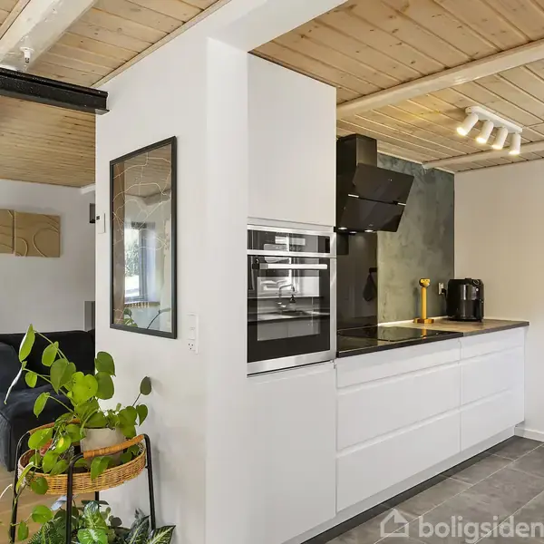 Moderne køkken med hvide skabe og sorte apparater. En trappe er ved siden af køkkenet, og en stue med en sort sofa er til venstre. Trægulv og træloft.