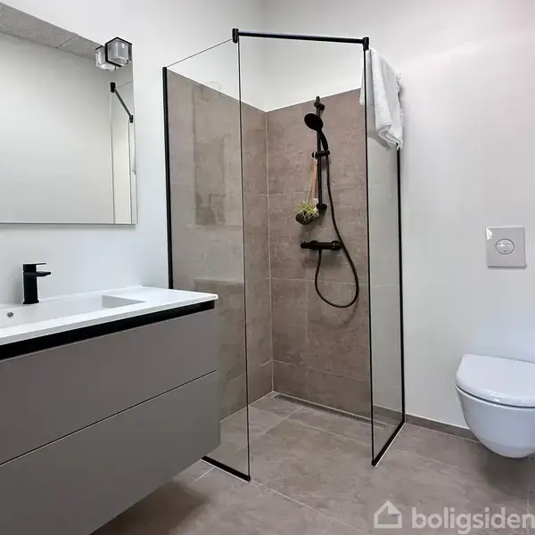 Badeværelse med sort brusekabine, moderne armaturer og glasvægge. Til venstre en vask med to skuffer og stor spejl. Til højre væghængt toilet. Enkel og moderne indretning.