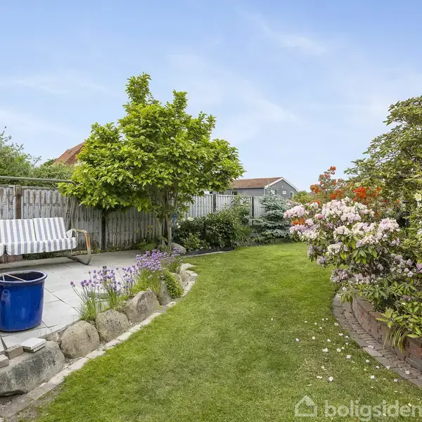 Træbord med blomsterbuket står i en hyggelig stue med sofa og spiseplads. Store vinduer slipper lys ind, der oplyser træloft og vægdekorationer.