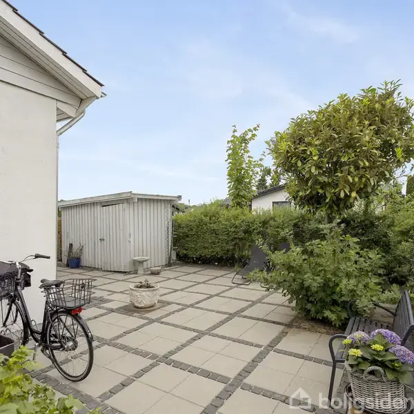En flisebelagt terrasse med en cykel i forgrunden. En træskur og en potteplante ses til venstre. Buske og et træ omkranser området under en klar himmel.