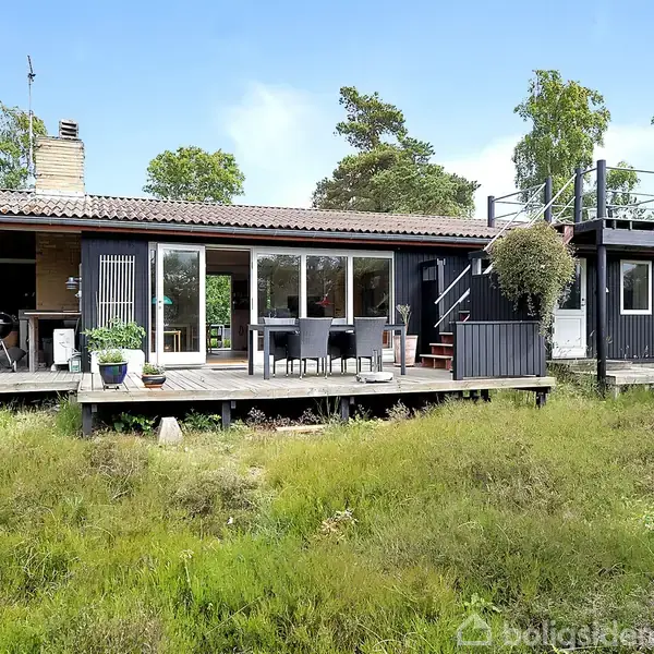 Huset, en træbeklædt bygning med store vinduer, har en terrasse med sofaer og grill. Det ligger i en skovklædt have med trampolin og træer omkring.