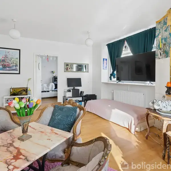 Stue med sofa og marmor bord, dekoreret med vase og tulipaner. Tv hænger på væggen ved vinduet. Hylde fyldt med pynt. PC i baggrunden. Seng ved siden af tv'et.