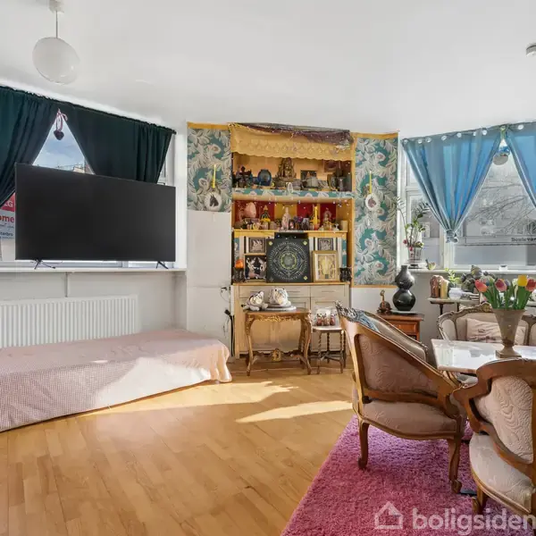 Stue med træmøbler og lavt bord dekoreret med blomster. TV hænger foran sofa ved vinduer med turkise gardiner. Indbygget reol i baggrunden fyldt med dekorative genstande.
