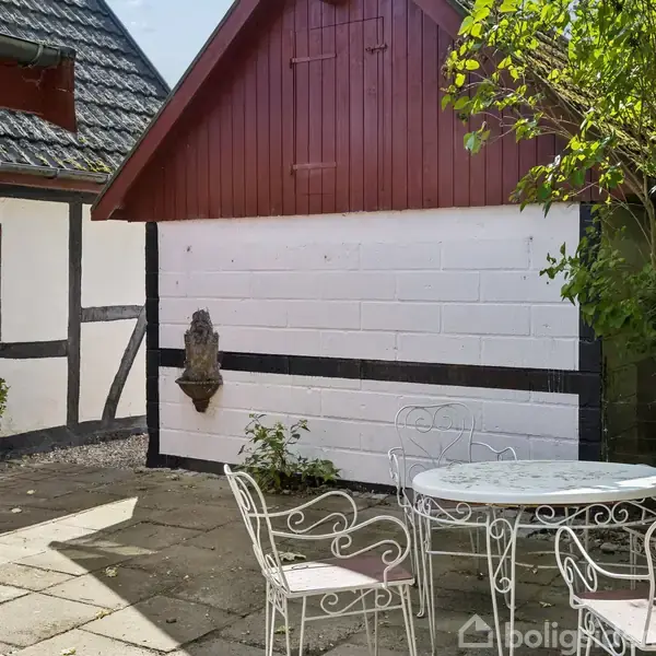 En hvid metalstol og bord står på en stenbelagt terrasse. En hvid bindingsværksmur med en lille skulptur i midten er bagved, omgivet af grønne buske og to bygninger.