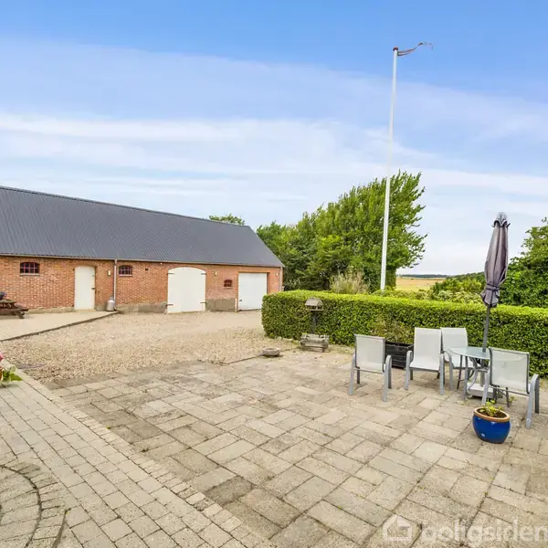 Terrasse med havemøbler står stille foran en rød murstensbygning med sort tag. Til venstre ses en lille stensætning, og baggrundens grønne buske grænser op til en åben mark.