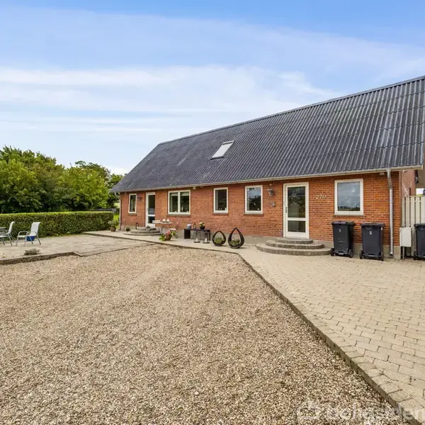 En rød murstensbygning med sort tag står stille. Omkring husets indkørsel ses flisebelægning og grus, mens træstol og træbord står i solbelyst have udenfor.