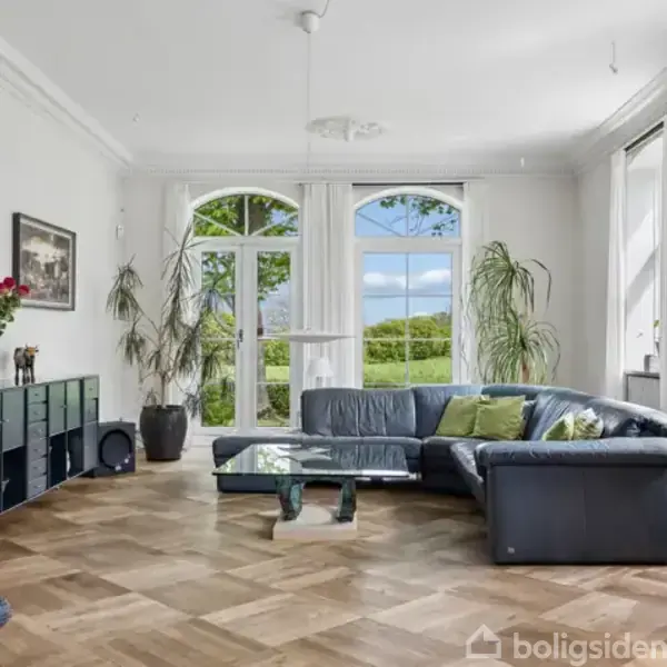 Sofa står ved siden af et stort glasvindue og glasdør med udsigt til træer og himmel. Stuen er lys med hvide vægge, gulv i parket, planter og et sort reolsystem.