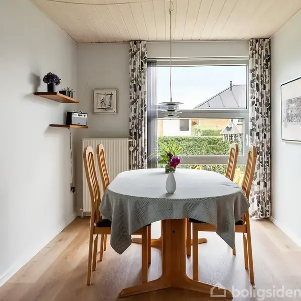 Et spisebord med fire stole står i et lyst køkken. Bordet er dækket med en dug, og der står en vase med blomster på det. Vinduet bagved har gardiner.