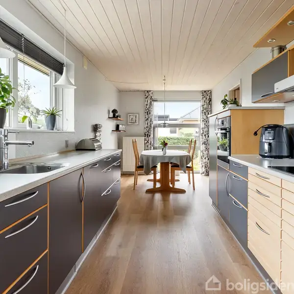 Køkken med mørke skuffer og hvide bordplader. Køkkenmaskiner står på bordet. I baggrunden er der et spisebord ved et vindue med udsigt og gardiner. Planter på vindueskarmen.