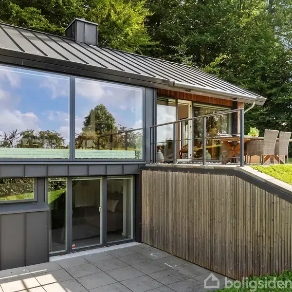 Moderne hus med store glaspartier på en hævet terrasse, omgivet af frodig græsplæne og træer. Spisebord og stole står på terrassen ved siden af glasvæggen.