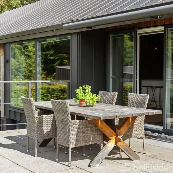 Et træbord med fire fletstole står på en terrasse. Det er omgivet af en moderne bygning med store vinduer og grønne omgivelser. En potteplante står på bordet.