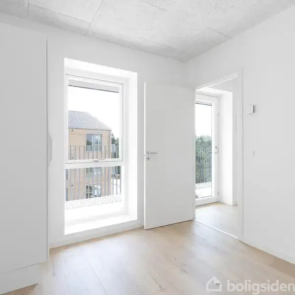 Et lyst rum med trægulv har en åben dør, der fører til en balkon med glasramme. Store vinduer giver udsigt til en bygning udenfor.