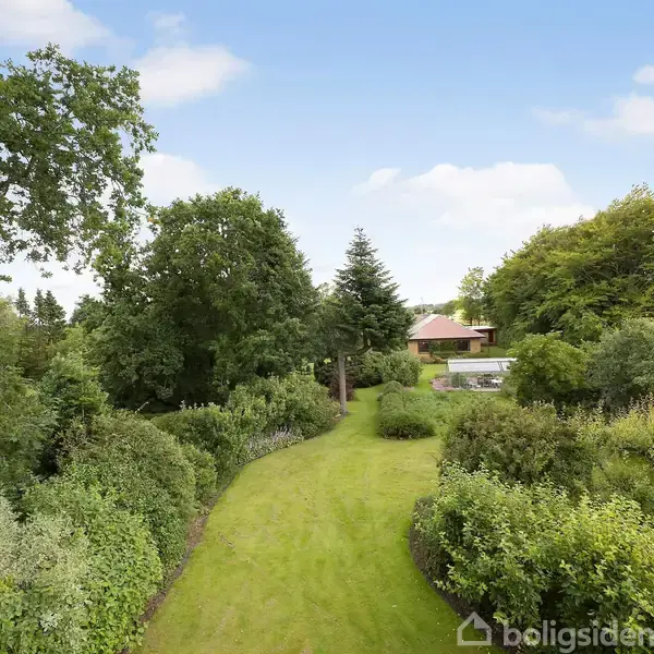 Grøn græsplæne strækker sig gennem en frodig have med store buske og træer, fører til en villa i baggrunden. Klart solskinsvejr med blå himmel og lette skyer.