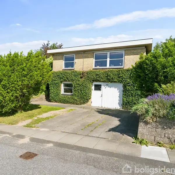 Et to-etagers hus dækket med klatreplanter, står stille foran en garage med en asfalteret indkørsel. Omgivet af buske og blomsterbede i et beboelseskvarter.