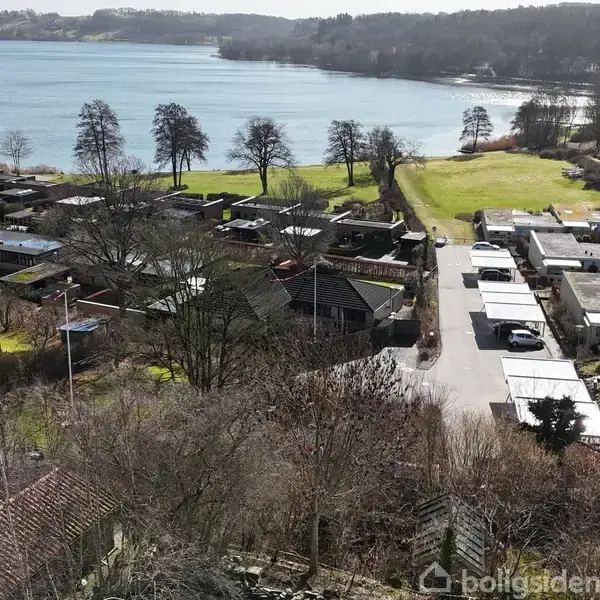 En række huse står stille langs en vej i en frodig, kuperet landskab. En sø breder sig i baggrunden, mens træer omkranser området.