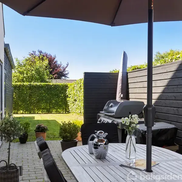 Parasol dækker spisebord med vase og karaffel, mens grillen står på terrassen. Omgivet af hæk og solbeskinnet have.