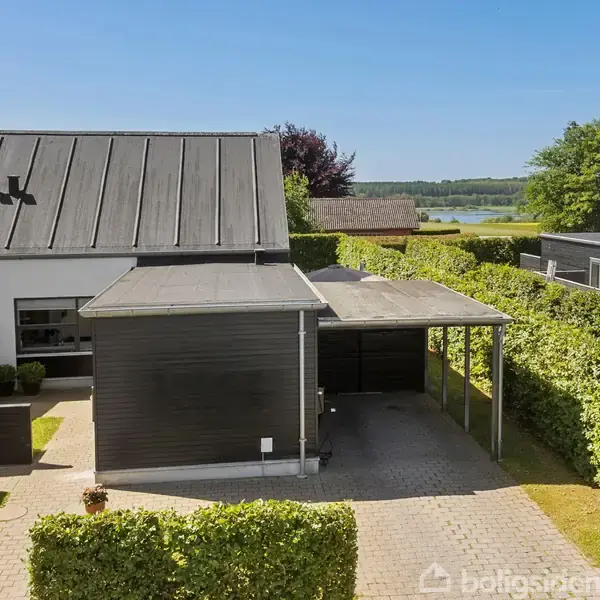 En moderne, sort-hvid bolig med fladt tag står stille i et velplejet haveområde. Den har en overdækket carport og er omgivet af grønne hække. En skov og sø kan ses i baggrunden.