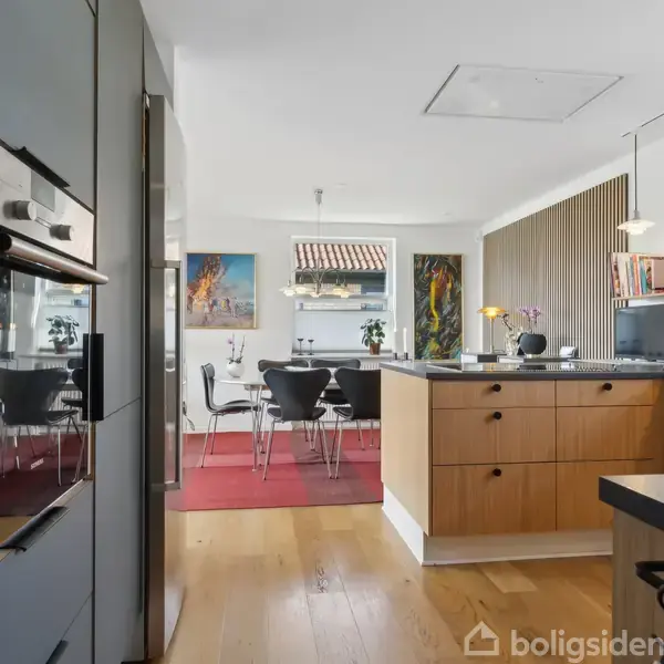 Køkken i moderne design med indbyggede apparater, træskabe og en ø. Til højre ses spiseområde med sort bord og stole, dekoreret med kunst på væggene og en rød tæppe.
