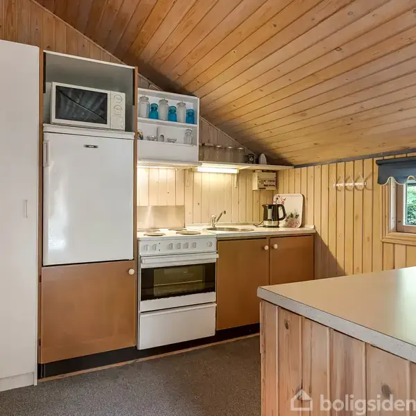 Et lille køkken med trævægge indeholder et køleskab, mikrobølgeovn, ovn og kogeplader. Over disken ses kopper og glas på hylder. Et vindue med blå gardiner tillader udsigt til grønne omgivelser.