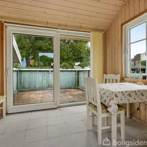 Træbeklædt rum med to stole og et tæppebelagt bord ved et vindue. Skydedøre åbner ud til en træterrasse med udsigt til grønne træer.