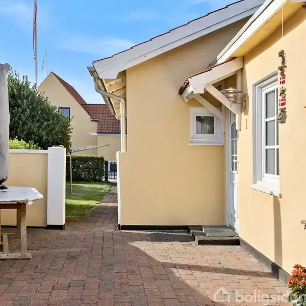 Et gult hus med rødt tag står i en solrig gårdhave. På en flisebelagt sti ses et bord med en sammenfoldet parasol. Blomster pryder hjørnet af huset.