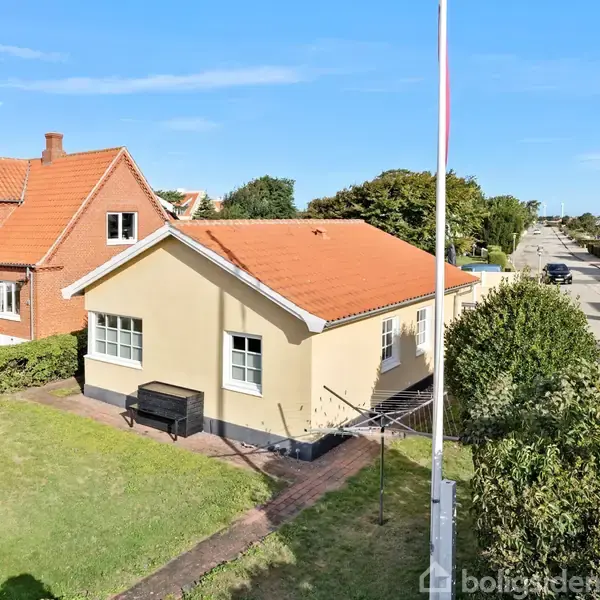 Et gult hus med rødt tag står på en grøn græsplæne ved siden af en vej. Der er en flagstang i forgrunden og buske omkranser grunden.