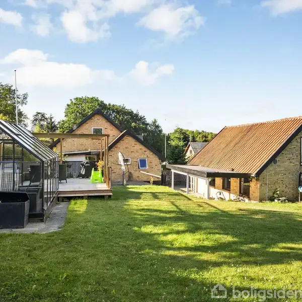 Væksthus ved siden af terrasse med legetøj i forhave; baggrund viser to murstenshuse med skrå tage og en trampolin ved siden af græsplæne under blå himmel.