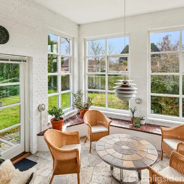 Et lyst rum med rattanmøbler omkring et rundt mosaikbord. Store vinduer giver udsigt til en frodig have. En moderne lampe hænger over bordet, omgivet af planter.