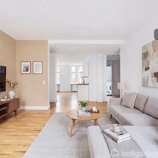 En moderne stue med en grå sofa og et rundt træbord. Et TV hænger på væggen, og en træskænk står under det. Vægkunst og planter tilføjer dekoration. Tekst: "Visualisering".