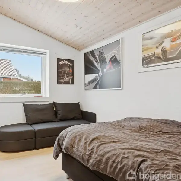 Soveværelse med en grå seng til højre og en sort sofa under et vindue til venstre. Væggene prydes af tre bilplakater.