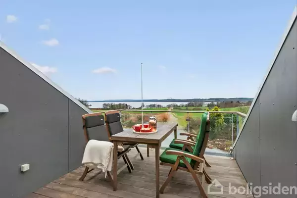 Et træbord med stole står på en terrasse med udsigt til en sø og grønne områder. Et tæppe hænger over en stoleryg ved siden af en tebakke.