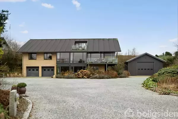 Hus med moderne design står i en gårdsplads, omgivet af grus og vegetation. Store vinduer og balkon er synlige, med tilhørende garage på højre side.