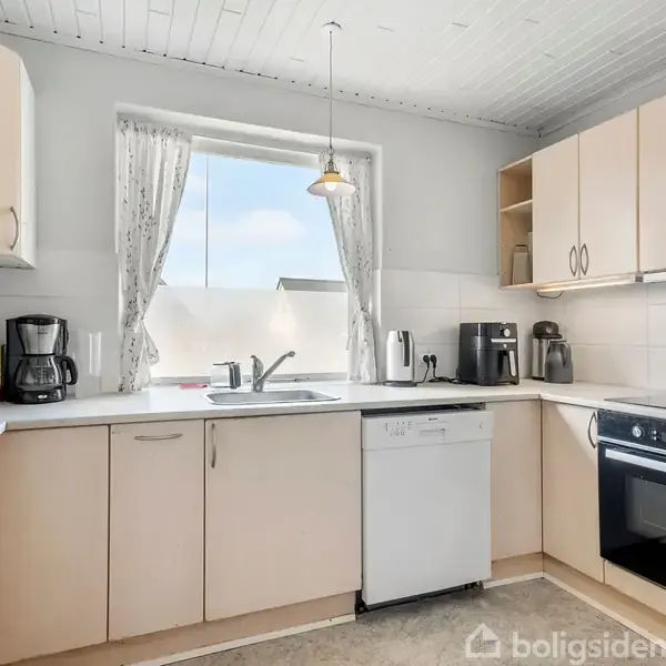 Et lyst køkken indeholder lysebrune skabe, opvaskemaskine, indbygget ovn og køkkenredskaber. En vindue med gardiner lader dagslys komme ind, hvilket skaber en lys atmosfære.