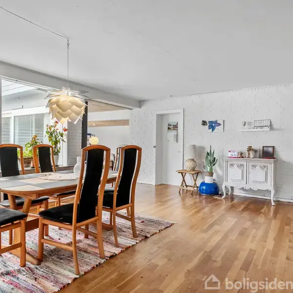 Spisebord med seks stole står på et farverigt tæppe. Over bordet hænger en stilfuld lampe. Rummet har lyse vægge, trægulv, store vinduer og et hvidt skab i hjørnet.