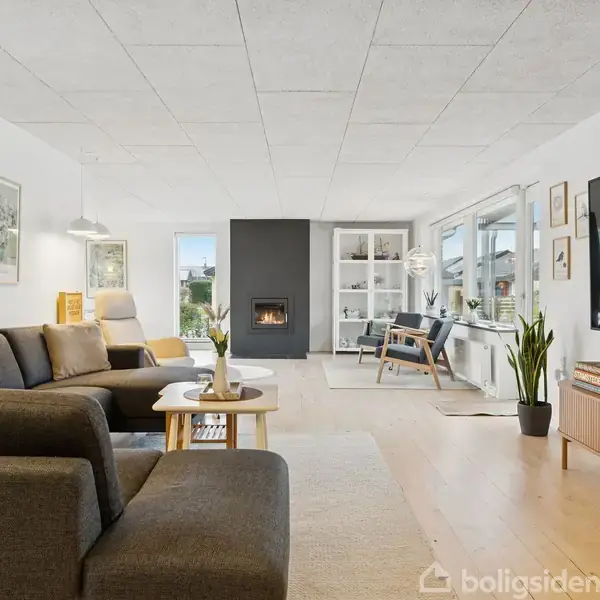 En stue har en mørk sofa med sofabord foran, indrammede billeder på væggen, en pejs i midten, reoler i hjørnet og store vinduer til højre.