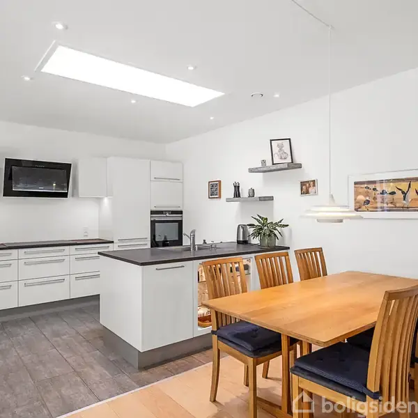 Køkken med hvidt køkkensæt og sorte bordplader, inkluderende indbyggede apparater; spisebord med træstole i forgrunden. Rummet er lyst med træ/laminatgulve og moderne indretningselementer.