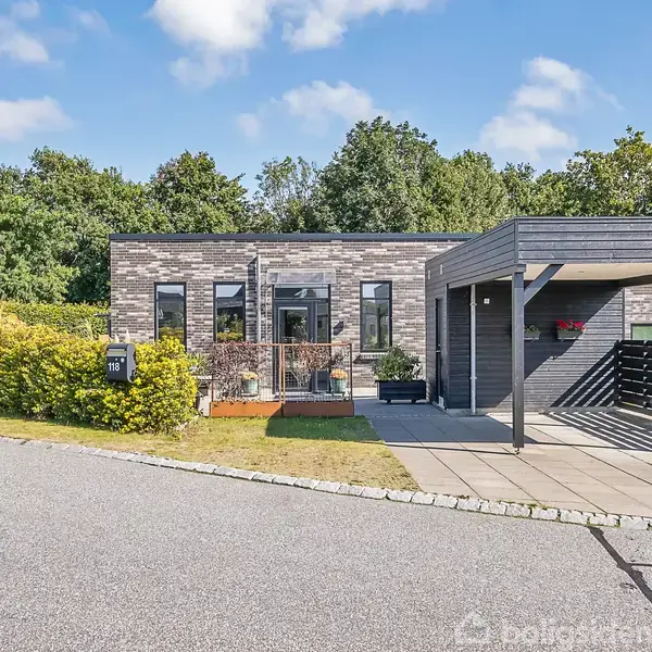 En moderne etplansvilla med fladt tag står stille ved en asfaltvej. Den har en tilhørende carport til højre. Omgivet af grøn buskads og træer i baggrunden.