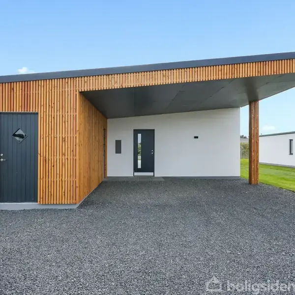 Moderne hus med træbeklædt overdækning står stille på en grusbelagt indkørsel, flankeret af en velholdt græsplæne og klar himmel i baggrunden.