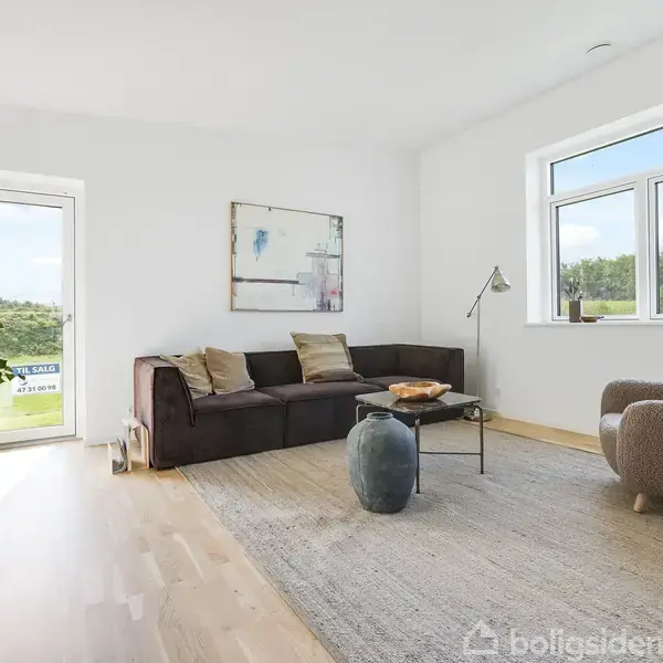 Sofa med puder står i et lyst rum ved siden af et sofabord med vase og stol. Store vinduer og en dør fører ud til en grønt område.