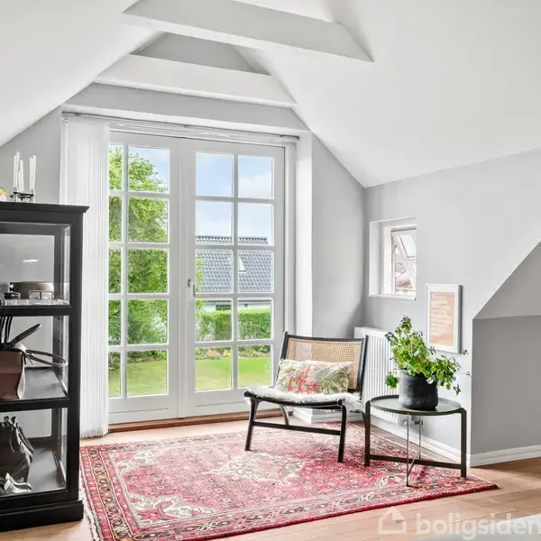 En lys stue med en fransk dør åbner mod haven. En sort reol står til venstre, en stol og et lille bord med en plante til højre på et rødt tæppe.