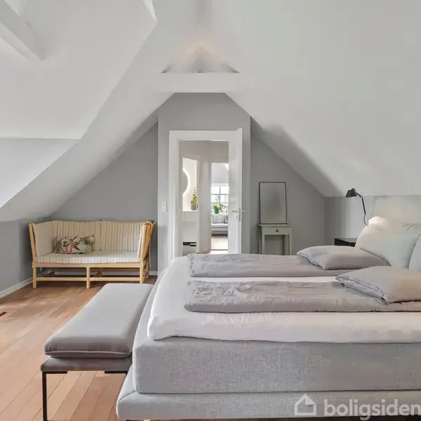 Soveværelset har en dobbeltseng placeret under et skråt loft, en bænk for enden af sengen, og en sofa ved siden af et vindue. Rummet er lyst og indbydende.